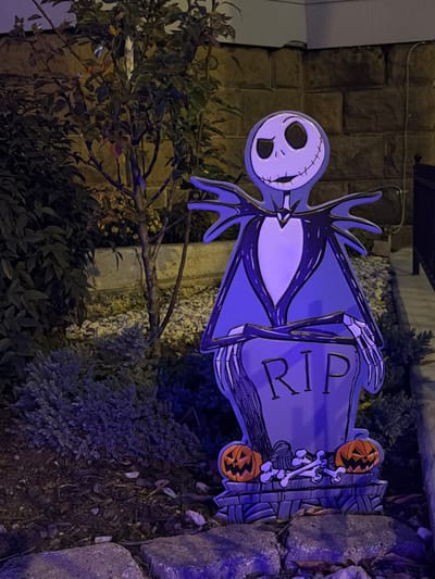 A Halloween yard decoration featuring Jack Skellington stands upright in a garden bed in Weehawken, United States. The cutout depicts Jack Skellington, dressed in his black pinstriped suit with a white face and stitched smile, holding his skeletal hands on a grey tombstone prop marked with "RIP." The base of the decoration includes two orange jack-o'-lanterns and scattered white bones. The scene is illuminated by artificial light, casting a prominent purple hue on the decoration and surrounding low-lying evergreen-like shrubs. Behind the decoration and to its left, a slender deciduous tree with sparse foliage and dense dark green shrubbery are visible. In the background, a wall constructed from large, rectangular light-colored stone blocks extends horizontally. To the far right, a dark railing is partially discernible. The immediate foreground shows several uneven grey stones, forming a garden border over dark earth or mulch. The overall setting is an outdoor, low-light environment at night.
