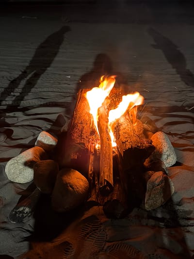 A bonfire blazes in the darkness, flames licking at the stacked logs. Rocks of varying sizes surround the fire pit. The ground appears to be textured, with what looks like ripples or grooves, possibly sand. A long, indistinct shadow stretches across the surface, indicating a presence near the fire. A footprint impression is present.