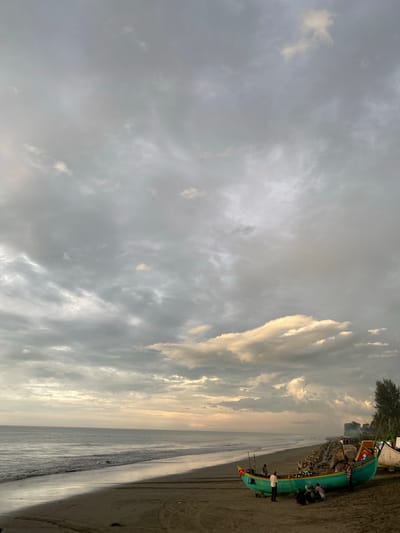 This image depicts a beach scene in Bateetimoh, Indonesia, under a cloudy sky. In the foreground, a brightly colored teal and yellow fishing boat rests on the sandy shore, with several individuals gathered around it. The boat is adorned with various decorations and appears to be partially loaded with yellow netting. Further down the beach, more people can be seen walking or standing, and a rocky breakwater stretches along the coastline. The ocean stretches to the horizon, with gentle waves lapping at the shore. The sky is filled with dramatic, overcast clouds, with patches of sunlight breaking through in the distance, suggesting either sunrise or sunset. Trees and foliage are visible on the right side of the image, indicating the presence of vegetation inland from the beach. Tire tracks are visible in the sand, suggesting vehicle activity on the beach.