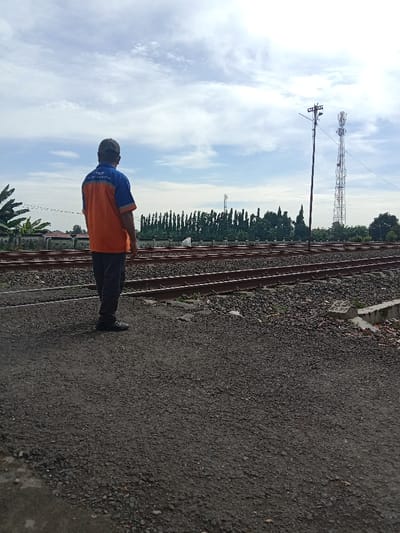 A man stands with his back to the viewer, wearing a blue and orange shirt and a cap, on a dark, gravelly surface. In the background are several sets of railroad tracks with greenery beyond. Two communication towers rise in the distance against a sky filled with white clouds. The scene is captured in natural light, suggesting a daytime setting.