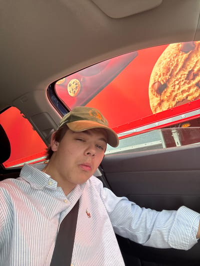 The image shows a young man, wearing a green baseball cap and a light blue and white striped button-down shirt, sitting in a car. The man has short, dark hair and appears to be looking directly at the camera. He is wearing a seatbelt. The car window is rolled down, revealing a red truck with a large advertisement on the side. The advertisement features a large chocolate chip cookie. The coordinates provided place this scene in the town of Cleckheaton, in the Kirklees metropolitan borough of West Yorkshire, England.