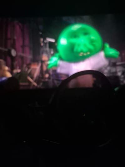 The image depicts a dimly lit scene, possibly indoors, with a prominent, glowing green object in the foreground, appearing somewhat spherical with indentations that suggest facial features or a stylized design. To the left of this object, a green, stylized hand or limb is visible, also glowing. Behind the green object, a blurred figure in light-colored clothing can be discerned. The background is dark and indistinct, with hints of what might be furniture or decorative elements, including some purplish hues. The right side of the image is almost entirely black, obscuring any details. A dark, rounded object, potentially a piece of equipment or a camera lens, is partially visible in the dark area, reflecting some light. The overall impression is of a somewhat surreal or artificial environment, possibly a display or an exhibit, given the glowing nature of the primary subjects. There is no discernible text, weather, or time of day information. The emotion conveyed is ambiguous due to the low light and blurriness.