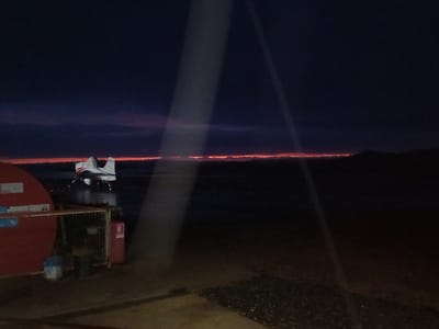 The image displays an outdoor scene at twilight or night in Santa Juana, Chile, characterized by low light conditions and a prominent horizon glow. In the midground, a white single-engine floatplane with red accents rests on a dark body of water, facing slightly towards the left. The aircraft features two visible pontoons. Across the distant horizon, a distinct, narrow band of intense orange-red light is present, likely indicating sunrise or sunset, above a silhouetted landmass. The sky above this band is dark blue to purple, transitioning to black.

In the immediate foreground, to the left, a portion of a large, curved red object, possibly a fuel tank or structure, is visible. Adjacent to it, a metal cage-like container holds various objects, including a blue barrel and a dark container. A red fire extinguisher is mounted nearby. The ground in the foreground is dark, appearing to be textured pavement or gravel. Two diagonal light streaks traverse the upper-left to mid-right portion of the image. No individuals are present in the scene.