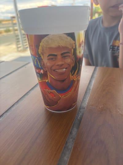 The image shows a close-up of a paper cup with a graphic of a person's face on it, placed on a wooden table. The person on the cup has blond hair and is smiling, with braces visible. The cup has a white rim and is red and yellow on the lower half. To the right of the cup, part of a person's torso and shoulder is visible, wearing a gray t-shirt with a logo that appears to be "PER". The background shows a blurred outdoor scene with a railing and a cloudy sky. This scene is located in Drazhevo, Bulgaria, at a McDonald's restaurant.