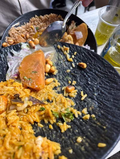 An overhead, close-up shot captures a vibrant Peruvian meal being served or eaten, likely in Jesús María, Peru. The scene is dominated by two dark, textured ceramic bowls filled with food, with a hand holding a metal spoon extending into the frame from the left.

The foreground bowl contains a rich, orange-yellow rice dish, likely *arroz con mariscos* (rice with seafood), identifiable by the cooked rice grains mixed with pieces of seafood (shrimp and possibly other shellfish/fish), green herbs (such as cilantro), and hints of a savory sauce. Scattered throughout this dish and on the rim of the bowl are toasted peanuts or *cancha* (Peruvian toasted corn).

Adjacent to the seafood rice, within the same bowl, is a section featuring thinly sliced red onions and a prominent, large, orange chunk of what appears to be cooked sweet potato (*camote*), often served with ceviche. More peanuts are visible near these components.

In the background, a second, slightly out-of-focus bowl holds a lighter brown, fluffy rice, possibly plain rice or a different rice preparation, also with scattered peanuts.

To the upper left, partially obscured, is a clear glass containing a bright yellow, effervescent liquid, consistent with a soft drink like Inca Kola or beer, and part of a bottle with a blue label is also visible, though no text is legible. The overall setting appears to be a casual dining experience, with the focus solely on the delicious food.