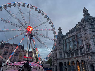 The image presents a striking urban landscape in Antwerp, Belgium, captured under an overcast sky, likely during dusk or evening. A large Ferris wheel, brilliantly illuminated with a pattern of red, white, and blue LED lights, dominates the left side of the frame. At its hub, a circular sign prominently displays a red 'A' logo and the text "THE VIEW ANTWERPEN," indicating its function as a scenic attraction in the city. The Ferris wheel features numerous enclosed gondolas.
To the right, a grand, ornate stone building stands tall, showcasing intricate architectural details, multiple domed towers, spires, and large arched windows. This impressive structure is Antwerp Centraal Station, with the text "CENTRAAL STATION" faintly visible on its lower facade. Its classical, historic architecture creates a stark contrast with the modern, brightly lit Ferris wheel.
In the background on the far left, another historic building can be seen with arched windows, partially obscured by the Ferris wheel's structure. A banner on this building clearly reads "ZOO ANTWERPEN," confirming the proximity of the city's zoo to this lively area. A few indistinct figures are visible at the base of the Ferris wheel, suggesting public activity. The overall impression is one of a vibrant city center where historic landmarks meet contemporary entertainment.