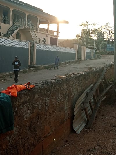A sunny street scene shows a multi-story building with an ornate fence and exterior staircase. Two people walk along the street in the distance, with a dog nearby. A low stone wall runs along the edge of the road, partially obscuring discarded items: laundry, a ladder, and a sheet of corrugated metal. The background features trees and another building with weathered towers. The overall impression is of a residential area with signs of everyday life.