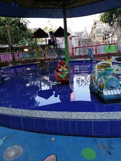 The image captures a public swimming pool area, likely a water playground, situated in Kebonsari, Indonesia. The pool itself is filled with deep blue water, reflecting the overhead canopy and surrounding trees. In the foreground, a small, colorful toy train with a blue and green color scheme floats on the water. To the left, a red and green character resembling an angry bird is also in the pool. The edge of the pool is lined with a mosaic of small, light-colored stones, transitioning to blue tiles. Beyond the pool, a metal fence with a red railing encloses the area, and beyond that, a sign reading "LOCK V" is visible. In the background, various structures and playground equipment, including slides and a blue dolphin statue, are present. The overall environment appears to be an outdoor recreational space with a festive and playful atmosphere, though the lighting suggests it might be late afternoon or early morning.