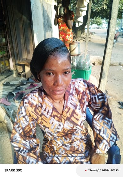 The image depicts an outdoor scene in the city of Garaku, Nigeria, under bright daylight. In the foreground, a young adult female with dark skin, styled black hair, and a slight smile looks directly at the camera. She wears a long-sleeved top or dress with a brown, orange, and white geometric pattern and a thin gold-colored necklace. Her skin shows a sheen, possibly from sweat or oil, with visible texture. She appears to be seated, with a blue armrest visible on her right.
Behind the foreground subject, to her left, a collection of various sandals and slippers are scattered on dusty ground near the base of a light-colored building with a wooden door. A wooden bench is attached to the building wall. To the right of the foreground subject, a green plastic crate holds numerous empty glass bottles, some bearing "Pepsi" branding. Next to the crate is a light-colored cylindrical container.
Further into the background, a second individual with dark skin, wearing a red and orange patterned dress, stands and holds a metallic cup to their mouth, appearing to drink. White fabric items hang near a tree with green foliage. The ground in the mid and background is unpaved and dusty, suggesting a street or pathway. A motorcycle is parked on this path, and part of a light-colored car is visible further in the distance. A light-colored animal, possibly a goat, is partially discernible on the ground behind the crate. A prominent wooden pole stands vertically on the right side of the frame.