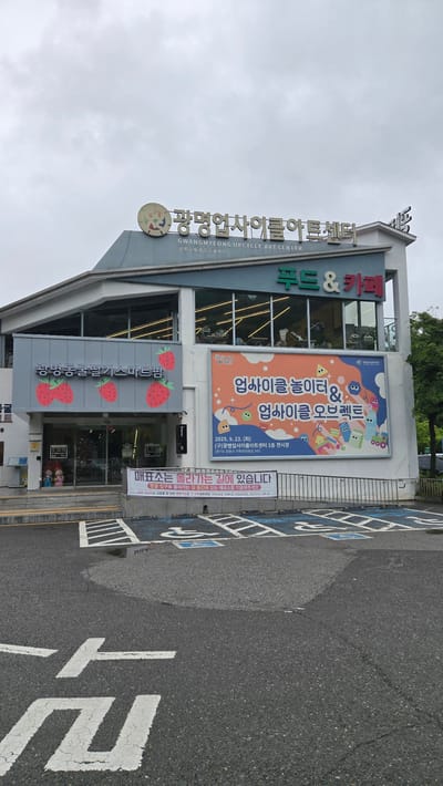This is a photograph taken outdoors in daylight, under an overcast sky, showcasing the exterior of the Gwangmyeong Upcycle Art Center in Gwangmyeong, South Korea. The building features a modern, multi-story design with large windows. A prominent vertical sign displays the Korean characters "광명업사이클아트센터" (Gwangmyeong Upcycle Art Center) in large, metallic gold letters, with smaller English and Chinese text below.
A large, colorful banner is displayed on the upper part of the building, advertising an event related to "Upcycle Playground & Upcycle Objects," scheduled for September 23, 2025, at the first-floor exhibition hall. The banner features cartoonish illustrations of various objects, some of which appear to be made from recycled materials.
Below the banner, the entrance area is decorated with large, stylized red strawberries. The name "광명동굴딸기스마트팜" (Gwangmyeong Cave Strawberry Smart Farm) is visible in Korean script on this section of the building.
Adjacent to the building is an asphalt road with designated parking spaces marked with blue paint and white lines. These parking spaces are unusually wide, and one of them appears to be a handicapped parking spot. In the foreground, large white Korean characters are painted on the road surface, forming part of a sign that reads "매표소는 올라가는 길에 있습니다" (The ticket office is on the way up). This indicates that the building is likely a tourist attraction or a venue with a ticketed entry.
While no people are clearly visible in the immediate foreground or in the parking lot, some figures can be seen through the glass doors of the building, suggesting that the venue is open and possibly in operation. The overall impression is of a community-focused arts and culture center with a unique theme of upcycling and potentially featuring local produce or attractions like the "strawberry smart farm."