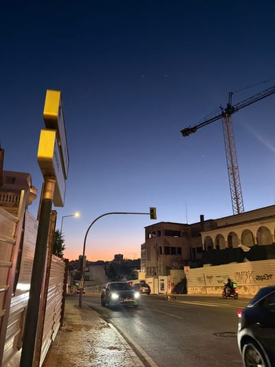 This image captures a twilight scene on a street in Cascais, Portugal. The sky transitions from a deep blue overhead to a warm orange hue near the horizon, indicating the time is either dawn or dusk. A large construction crane dominates the upper right portion of the frame, silhouetted against the sky.
In the foreground, a sidewalk runs along the left side of the image, bordered by a corrugated metal construction barrier. A yellow traffic signal with a green light is visible above the street. On the street, a dark-colored car is driving, its headlights illuminated. Further down the road, a scooter rider wearing a green delivery bag is visible.
The background features several buildings, some appearing to be under construction or renovation, with exposed structures and scaffolding. The architectural style includes arches and balconies, consistent with Portuguese design. The street is paved and appears wet, reflecting the streetlights. The overall impression is a busy urban environment at the end of the day.