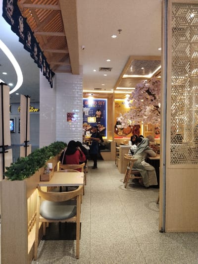 The image shows the interior of a restaurant in Telukjambe, Indonesia, located at coordinates -6.301 latitude and 107.278 longitude. The restaurant is decorated in a modern Japanese style, with wooden floors and walls, and a large cherry blossom tree in the background. There are three people seated at a table, and one person standing near the table, presumably waiting for their order. All are dressed in contemporary clothing. The tables and chairs are made of light wood, and the walls are covered in white tiles. There are potted plants along the walls, creating a relaxing atmosphere.  The scene appears to be taken during the afternoon.