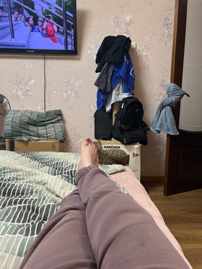 The image depicts a domestic indoor scene, possibly in Tula, Russia, focusing on a person's legs and feet resting on a bed. The bed is covered with a comforter featuring a striped pattern in muted green and grey tones. To the right of the bed, a person's legs, clad in dark purple sweatpants, are visible. A cat, possibly a tabby, is curled up on a box near the foot of the bed.
The background reveals a wall adorned with floral wallpaper in pale pink and white hues. Several items are hung on the wall, including clothing items like a black jacket, a blue towel, and striped fabric, as well as a white electrical outlet. A large black backpack rests on top of a white box labeled "KÄRCHER". A television screen is mounted on the wall in the upper left corner, displaying a scene with several people. To the right, a wooden door frame is visible, with a blue towel draped over the handle. The floor appears to be wooden. The scene suggests a casual, relaxed atmosphere within a residential setting.