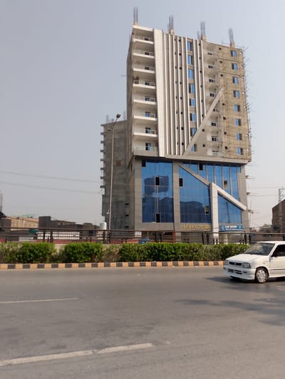 In Peshawar City Tehsil, Pakistan, a tall, modern building under construction, identified as "SAI AFI TOWER," dominates the scene. The tower, with its white facade and reflective blue windows, is partially clad in scaffolding on its right side, indicating ongoing construction work. Below the tower, a white car is driving on a paved road. The road is lined with a low wall and dense green bushes. In the background, other buildings and a lamppost are visible, under a clear sky.