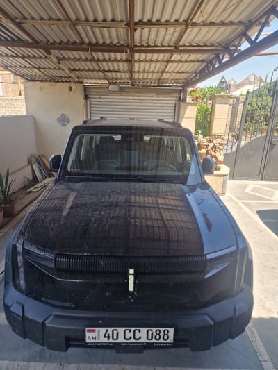 A black SUV is parked under a corrugated metal roofed carport in Nor Kharberd, Armenia. The front of the vehicle is facing the camera and is covered in a thin layer of dust. The license plate reads "40 CC 088" and has Armenian characters. Behind the vehicle, a closed garage door is visible. To the right of the garage, a decorative metal gate with ornate scrolls is open, revealing a glimpse of greenery and stone structures beyond. The ground is paved with concrete tiles. A potted plant with green leaves is situated to the left of the vehicle. The overall environment appears to be a residential or semi-industrial area with a dry, sunny climate.