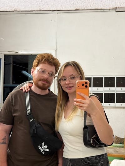 A young couple is reflected in a mirror, likely taking a selfie. The woman, with blonde hair and glasses, holds an orange phone. She wears a white top and a black shoulder bag. The man, with red hair and a beard, wears glasses and a brown t-shirt, with a black Adidas fanny pack across his chest. A tattoo is visible on his arm. Behind them is a white wall with square black and white panels. The reflection also shows part of a window and greenery. The location appears to be indoors. This is in Rehovot, Israel, given the prompt.