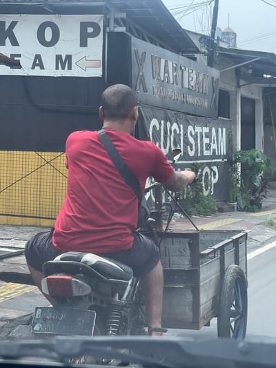 The image shows a man riding a motorcycle with a sidecar attached. He is wearing a red t-shirt and dark shorts. The motorcycle is pulling a metal cart. In the background, there is a building with signs that read "KOP" and "WARTEAM X". Another sign says "CUCI STEAM". The scene appears to be on a street in a city, possibly South Tangerang, Indonesia, given the license plate format (B 5556 Jf) which is typical for Jakarta and surrounding areas. The environment suggests an urban setting with buildings and a road. The lighting is overcast, indicating a cloudy day.