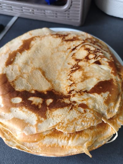 A stack of golden-brown crepes rests on a white plate, which is placed on a dark gray countertop. The crepes exhibit irregular browning patterns, with some areas darker than others, indicating they were likely cooked on a stovetop. The texture appears slightly porous. In the background, a portion of a brown and white striped storage container is visible, also on the countertop. The scene is lit from above, casting soft shadows. There are no people or animals present in the image. The location in Brodi, Latvia, is not discernible from the visual information provided.