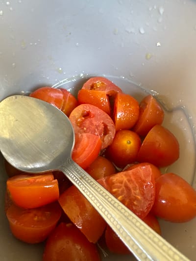The scene depicts a quantity of red cherry or small plum tomatoes, predominantly cut in halves or quarters, resting within a light grey bowl or container. A silver-colored metal spoon with a decorative patterned handle is positioned diagonally across the tomatoes, extending from the lower left towards the upper right of the frame, partially submerged among them. The inner surface of the container shows droplets of clear liquid, consistent with moisture from the tomatoes or washing. The tomatoes themselves appear fresh, displaying their internal structure including seeds and pulp. The setting is identified as Invergowrie, United Kingdom. No individuals or active interactions are visible within the frame.