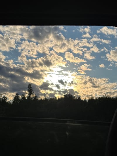This image depicts a view through a window, likely from a vehicle, looking out at a landscape at sunset. The upper portion of the image is dominated by a sky filled with a mixture of white and gray clouds, with the sun breaking through in the center, casting a warm, golden light. The lower portion of the image shows a dark silhouette of a dense line of trees, indicative of a forest or wooded area. Below the trees, a faint horizontal line suggests a road or barrier, and the foreground is dark, obscuring any details of the ground or immediate surroundings. The scene is consistent with the natural environment found in and around the city of Makaryev, Russia, a region known for its extensive forests and picturesque sunsets over the Volga River and its tributaries. No people or specific objects are discernible in the image, only the natural elements of the sky and treeline.