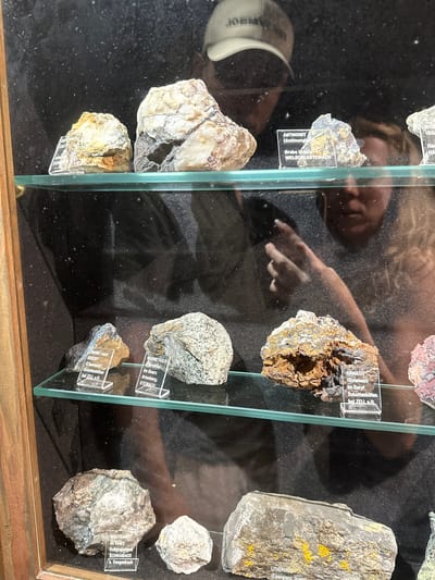 This media file shows a display case containing various mineral samples, likely in a museum or a collector's exhibit in Schonach im Schwarzwald, Germany. The display case has a wooden frame and glass shelves, with several minerals arranged neatly on a dark background. Each mineral specimen has a small label in front of it, identifying the mineral and its origin.
Visible text on the labels includes: "ANTIMONIT (Antimong Grube Ursula WELSCHENSTEINACH", "LIMONIT nach SIDERIT (Eisenspat) Schottenhöfen bel ZELL a.H.", "HAMATIT (Rotelsen) Im Baryt Schottenhöfen bel ZELL a.H.", "MAGNE TKIESS (Pyrrhotin) Im Quarz Artenberg. STEINACH", "WOLFRAMIT Im Quarz Roßgrabeneck SCHWAIBACH b. Gengenbach", and "LITHOPHYSE (Elsenglanz, Quarz)". The reflection on the glass reveals a person, likely a visitor, looking at the exhibit. The person's hands are visible, suggesting they might be pointing at a specimen or interacting with the display in some way. The lighting suggests an indoor setting, possibly during the day. The overall scene conveys an educational or recreational context related to geology and mineralogy.