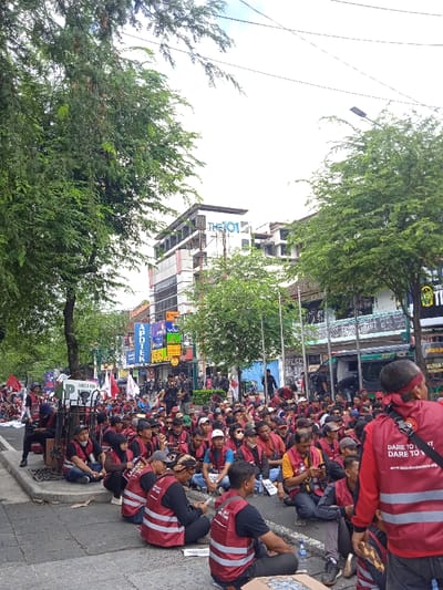 The image shows a street scene in Yogyakarta, Indonesia, taken at the coordinates -7.784 latitude and 110.367 longitude. A group of people, mostly men, are seated on the sidewalk in front of a building, many wearing red vests with the words "Dare to Fight for Justice". They are all facing the camera, some holding signs, banners, and flags. Several individuals are standing behind the seated group, observing the scene. The buildings in the background appear to be commercial, with signage and windows. There are trees on either side of the street, and power lines are visible above. The overall scene suggests a gathering of people engaged in a peaceful protest or demonstration.