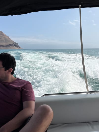 This media file shows a person on a boat sailing on the ocean near Callao, Peru. The person, with dark hair and wearing a maroon t-shirt, is partially visible, leaning forward with their head down. The boat is moving, creating a significant wake of white water. The scene is bright, suggesting it is daytime. The background includes a clear blue sky with some scattered clouds, and a rugged, rocky coastline is visible on the left side of the image. The overall impression is one of a leisurely boat trip on a pleasant day, with the focus on the motion of the boat and the ocean. There is no visible text in the image.
