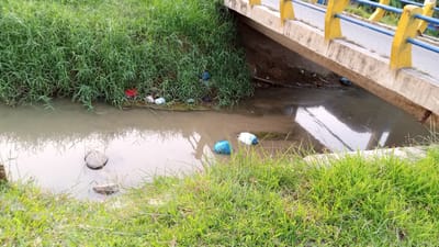 This media file depicts a polluted stream flowing under a bridge. The stream is murky and filled with debris, including plastic bags and other refuse. The banks of the stream are overgrown with tall green grass. The bridge, painted yellow and blue, spans across the water. The scene appears to be an outdoor environment, possibly in a rural or semi-urban area, given the presence of the bridge and the natural vegetation. There are no people visible in the image, and the overall mood is one of neglect and environmental degradation. The time of day appears to be daytime, with natural light illuminating the scene. There is no visible text in the image. The location context is Buketteukuh, Indonesia, which suggests this is a view of a waterway in that specific region.
