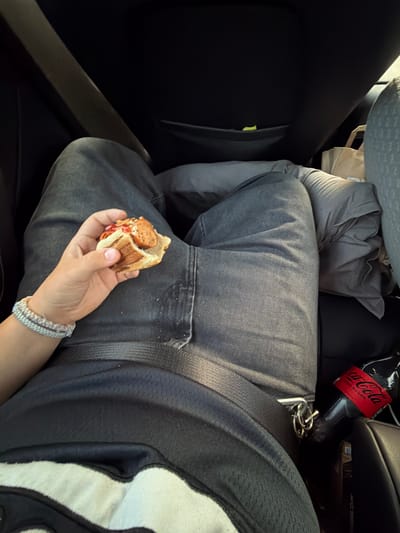A person's lap and arm are visible inside a vehicle. The person is wearing dark wash jeans and a black t-shirt. They are holding a sandwich made with a bun and a sausage in their left hand, which also has a bracelet on the wrist. A seatbelt is fastened across their lap. To the right, a bottle of Coca-Cola with a red label is visible, along with a set of keys attached to the seatbelt buckle. The background consists of car interior elements, including dark seats, a car seat pocket with a yellow object, and a crumpled grey pillow or blanket. The scene is consistent with being inside a car, potentially in the city of Hogstorp, Sweden, though no direct visual cues confirm this specific location.