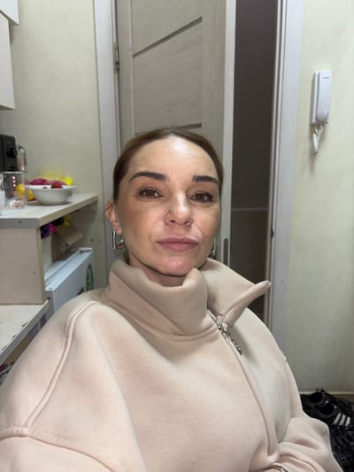A woman is looking at the camera. She is wearing a tan hooded sweatshirt with a zipper. She has brown hair pulled back and brown eyes. She is wearing silver earrings. She appears to be inside a building, possibly a home, as there is a door in the background and a shelf with items on it. It is not possible to verify the location in Novoe Devyatkino, Russia from the image.
