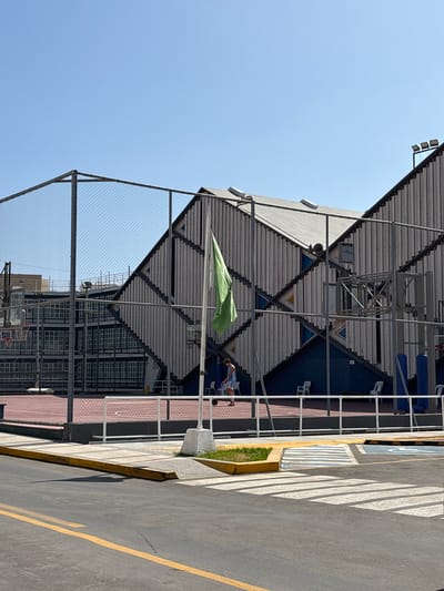 The image shows an outdoor basketball court with a modern, angular building in the background. A green flag is hanging from a pole in the foreground. A person is visible on the court, dressed in athletic wear and appearing to be playing basketball. The court is fenced with chain-link fencing. The building features a distinctive design with diagonal lines and corrugated metal siding. In the lower part of the image, a street with yellow lane markings and a pedestrian crosswalk is visible. The sky is clear and blue, suggesting a sunny day. The scene appears to be a public space in Chorrillos, Peru, likely an educational institution or recreational facility.