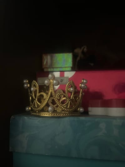 A gold crown adorned with pearls sits on a teal-colored box. Behind the crown, stacked boxes of red and green are visible. The background is dark and out of focus. The image is a close-up shot of the crown. There are no people present in the image, and no actions or interactions are depicted. The environment appears to be indoors, with the objects placed on a surface. The location in the city of San Antonio, Venezuela is not discernible from the image.