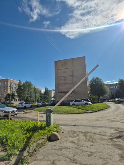 This is a sunny, daytime, outdoor, eye-level shot of a residential area in Petrozavodsk, Russia. The scene is dominated by a large, light-brown, brick apartment building on the right, with a white barrier arm extending diagonally from a green pillar to the left, angled upwards towards the building. The barrier is open and appears to be a traffic control device for a road that curves to the right. The road surface is asphalt with visible wear and tear, and patches of grass grow on the sides.

Several cars are parked along the street and on the grassy areas. These include a silver sedan in the foreground on the left, a white sedan, a light-colored SUV, and a dark sedan further down the road. To the left of the main brick building is a multi-story apartment building with many windows and balconies. A line of green trees separates the two buildings.

The sky is a bright, clear blue with some wispy white clouds and a prominent lens flare in the upper right corner, indicating the sun is high. Two faint contrails are visible in the sky. A light-colored, spherical object, possibly a decorative garden ornament or a fallen piece of equipment, sits on the grass near the barrier. The image appears to be taken in a suburban or urban residential district.