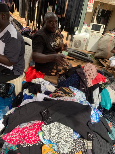 The image depicts an outdoor market scene in Nyanya, Nigeria, with two men present. In the foreground, a large pile of assorted clothing is displayed, featuring various patterns and colors including floral prints, polka dots, and solid black fabric. One man, wearing a black t-shirt and a patterned skirt, is seated on a wooden plank and gesturing with his hands. To his left, another man, wearing a black and white shirt and a black skullcap, is seated on a white plastic chair. Behind the clothing pile, several items of clothing hang on display racks. In the background, a white air conditioning unit, a refrigerator, and a microwave are visible, suggesting a mix of goods being sold. A large burlap sack is also present in the background, along with a sign that reads "OS." The ground appears to be dirt.
