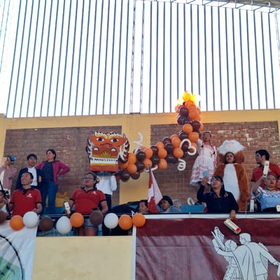 The image shows a group of people posing for a photo in front of a brick wall adorned with balloons and decorations. They appear to be part of a celebration or event, possibly a cultural or historical commemoration. A banner with silhouette figures and an illustration of a mask suggests the event is linked to Peruvian traditions.

The location is in Miraflores, Peru. The presence of Peruvian symbols on the banner, the type of brickwork, the architectural style, and local customs all point to Miraflores as the likely location.