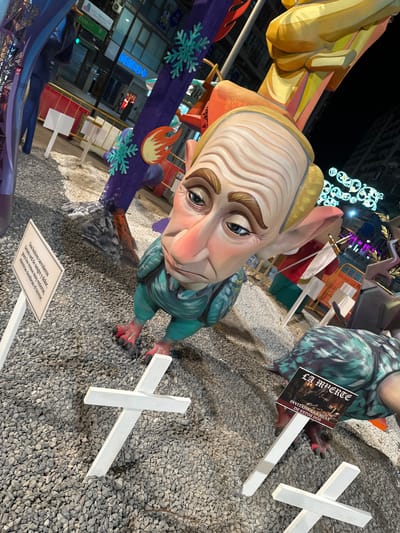 This is a medium shot, angled view of a large, elaborate effigy, likely part of a festival or exhibition. The central subject is a grotesque, cartoonish figure with exaggerated features, including a large head, pointed ears, and a prominent nose. The figure is painted in muted tones of beige, pink, and teal. It has large, expressive blue eyes and dark eyebrows. The figure is wearing a teal-colored garment and has red-clawed feet. It appears to be sitting or reclining on a bed of small grey pebbles.
To the left of the main figure, parts of another, larger, yellow and orange structure are visible, possibly another part of the same exhibit. A purple banner with snowflake and flame-like decorations is draped across the scene. In the background, city buildings and lights are visible, suggesting an urban setting, illuminated by artificial light, indicating it is nighttime or late evening.
Several white crosses are placed in the gravel around the figure, some upright, and one lying on its side. A small sign with text in Spanish is placed in front of the figure, and another sign with text and an image of skeletons is propped up near the figure's feet. The text on the signs, though partially obscured and angled, appears to be thematic and possibly narrative, relating to death or morbid themes, given the imagery and the phrase "LA MUERTE" (Death). The presence of such detailed, large-scale effigies and thematic signage points towards a festival like "Las Fallas" common in Valencia and Alicante, Spain. The overall mood is somewhat unsettling due to the figure's design and the thematic elements.