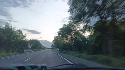 The image depicts a view from inside a moving vehicle on a two-lane road. On the left side of the road, there is a grassy verge followed by trees and what appears to be a bridge or overpass structure. The right side of the road is bordered by trees, some of which are blurred suggesting motion. The sky is overcast with light visible near the horizon, possibly indicating sunrise or sunset. The vehicle's dashboard and windshield are visible in the foreground. The location, Sokolovo, Bulgaria, is not discernible from the image.