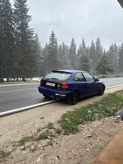 A dark blue Honda Civic hatchback is parked on the shoulder of a wet, two-lane asphalt road in a snowy, forested landscape in Pobit Kamak, Bulgaria. The car, viewed from its rear-left, features black steel wheels. The sky is overcast, and a light dusting of snow or frost covers the coniferous trees that line the road, as well as patches of grass on the verge.
Visible text on the car includes the Bulgarian license plate "PB 7300 PC" (indicating Plovdiv region), a "CIVIC" emblem, and a white sticker on the rear bumper that reads "DON'T HATE." Another small sticker is partially visible on the rear driver's side window.
The road has clear white and yellow line markings. A metal guardrail runs along the side opposite the car, separating the road from the dense forest. The shoulder consists of dirt and gravel, with some scattered debris and a black corrugated pipe visible on the far right. The scene depicts a cold, wintery or early spring day in a rural, possibly mountainous, setting. No people are visible.