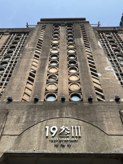 The photograph shows the exterior of the 1933 Shanghai building located in the Hongkou District of Shanghai, China. The building's facade is constructed of gray concrete. The design features alternating rows of circular openings and diagonal bars. The building's name is inscribed in stylized font: "1933 SHANGHAI." The sky is partially visible in the upper left corner. There are six black lights, three on each side, vertically positioned.