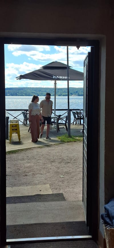 Two individuals, a woman and a man, stand on a wooden deck overlooking a body of water. The woman, on the left, is wearing a white lacy top and wide-legged brown pants. The man, on the right, is wearing a light-colored short-sleeved button-up shirt and dark shorts. They appear to be in conversation. Behind them, a large black umbrella with the logo "KARHU" is visible, along with several tables and chairs. The water stretches to a tree-lined horizon under a partly cloudy sky. The foreground shows a gravel path leading to a doorway, with a yellow stool stacked to the left of the deck. This scene is likely at a waterfront establishment in Lahti, Finland, potentially a restaurant or cafe.