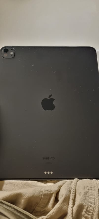 The image shows the rear view of a dark gray iPad Pro. The Apple logo is centrally located. Below the logo, "iPad Pro" is printed, followed by smaller text and three white dots. The top left corner of the iPad shows a camera array. The iPad is lying on a light brown, textured fabric surface. The background is a plain, light-colored wall. The image does not provide any information about the city of Başakşehir, Turkey.