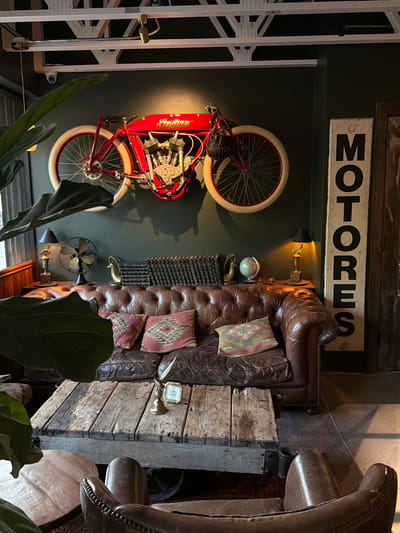 The image presents an interior design scheme with a vintage or industrial aesthetic, photographed at an angle.
The main elements include:
- A vintage-style motorcycle mounted on a dark-colored wall. The motorcycle is predominantly red with white rims and features the word "Indian" in gold lettering.
- Above the motorcycle, a sign with the word "MOTORES" in bold black letters.
- A leather couch with tufted cushions, adorned with decorative pillows.
- A rustic, wooden structure adjacent to the couch, which seems to serve as a side table or decorative element.
- Additional decorative items such as a globe, a small fan, books, and wall lamps are also present.
- There are indications of indoor plants in the foreground, obscuring part of the view.
The image suggests a bar, a club, or a restaurant in the city of West Hollywood, United States of America.