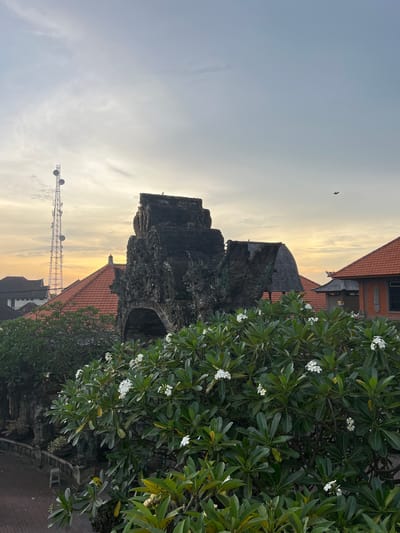 The image depicts a tall, ornate, dark-colored stone structure, possibly a gate or temple tower, against a pale sky with a yellowish-orange gradient. A bird is visible in the upper left. A blooming tree with green leaves and white flowers is present on the right side of the frame, partially obscuring the structure. Red-tiled roofs of buildings are seen in the background. In the lower left, a communications tower is visible against the sky. The location is likely Celuk, Indonesia, given the traditional architectural style.