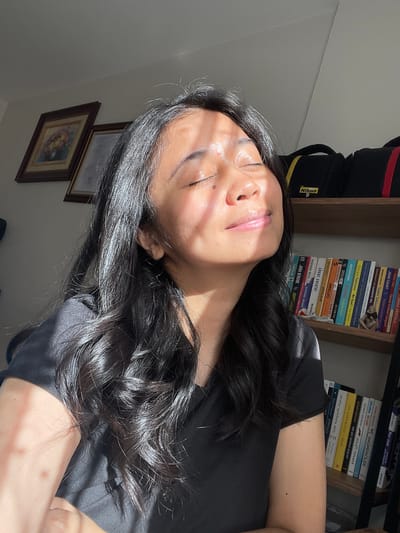A medium shot captures a young woman with long, dark, wavy hair in a close-up portrait. She is wearing a black t-shirt and her eyes are closed as she basks in the sunlight, which casts dappled shadows across her face and hair. The background reveals a bookshelf filled with books, some of which are visible on the right side of the image. Above the bookshelf, a shelf holds two black camera bags, one with a yellow and red stripe and the other with just a red stripe, and a Nikon logo is visible on the yellow-striped bag. On the left wall, two framed pictures hang, one depicting a floral arrangement and the other a lighter-colored picture. The lighting suggests the scene takes place indoors, likely in an apartment or home, and the presence of books and camera equipment implies a personal space within the city of Başakşehir, Turkey.