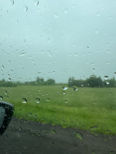 The image depicts a view through a rain-streaked window, likely of a vehicle, looking out onto a green, grassy field bordered by trees in the distance. The sky is overcast and a light gray. The foreground is dominated by numerous water droplets on the glass, distorting the view of the exterior landscape. A portion of a black car exterior, possibly a side mirror or part of the door frame, is visible in the lower left corner. The scene suggests an outdoor environment with inclement weather. No people or specific landmarks are identifiable to pinpoint a location within Arkhangelskoye, Russia.
