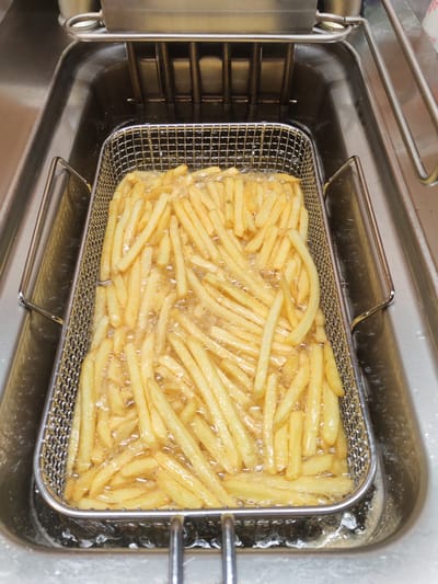 This image shows a close-up, overhead view of a metal basket filled with golden-brown french fries being deep-fried in hot oil within a commercial deep fryer. The basket is rectangular with a mesh bottom and sides, and has handles on each end. The french fries are cut into uniform sticks and are submerged in bubbling oil, indicating they are actively cooking. The deep fryer itself appears to be made of stainless steel and is part of a larger kitchen appliance. The surrounding environment suggests a commercial kitchen setting, likely a restaurant or fast-food establishment. There is no indication of the city of Hilversum, Netherlands, in this image.