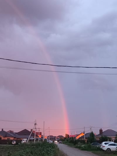 This is a photograph of a residential street in Ivanovka, Russia, at dusk. The sky is a dramatic mix of gray clouds and the soft, diffused colors of a sunset, with a prominent rainbow arcing across the upper portion of the frame. The rainbow, predominantly pink and red, creates a striking contrast against the muted sky.
The scene depicts a street lined with houses, some with dark tiled roofs and others with reddish-brown ones. A few vehicles are visible, including a white van and a white car parked on the side of the road, and cars driving down the street, their headlights illuminated. Power lines stretch horizontally across the sky, a common sight in residential areas. The foreground is filled with lush green vegetation, suggesting a rural or semi-rural setting. The overall mood is serene and atmospheric, with the colors of the sunset and the rainbow adding a touch of natural beauty to the ordinary scene. The time of day is clearly evening, as indicated by the sunset colors and the illuminated headlights.