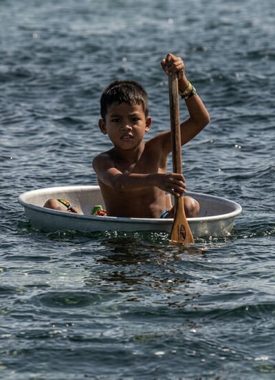 A young boy, appearing to be of Southeast Asian descent, is seated in a small, round, silver metal basin that is floating on dark blue water. He is shirtless and has dark hair. The boy holds a long, wooden oar, using it to propel himself through the water. His hands are on the oar, with his left hand higher than his right. He looks directly at the camera with a neutral expression.

Inside the basin with the boy, there are some colorful objects, including what appears to be a yellow toy and part of a brightly colored shoe or sandal. The basin is partially submerged in the water, indicating it is not a typical boat. The water is choppy, suggesting a slight breeze or movement. The sunlight is strong, casting reflections on the water's surface, and the overall lighting suggests it is daytime, likely midday.

The scene is set on a body of water, which could be a river, lake, or the sea. The backdrop is a blurred expanse of water, giving no specific indication of land. The boy's attire, or lack thereof, and the unconventional vessel suggest a possible rural or coastal setting. The overall impression is one of resourcefulness and a child navigating his environment.

There is visible text on the wooden oar, but it is too small and blurred to be deciphered.

The provided location context indicates the scene is in Soutr Nikom, Cambodia.