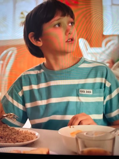 A young boy with dark hair is looking to his right with his mouth slightly open. He is wearing a teal and white striped t-shirt with a small pocket on the left side of the chest that has the words "COOL DUDE" embroidered in white. He is seated at a table with a plate of brown food in front of him, and a fork in his right hand. The background is blurred and appears to be a dining area with chairs and possibly other people. The image is a close-up shot focusing on the boy and his shirt. The scene is not identifiable as being in Vangaži, Latvia.