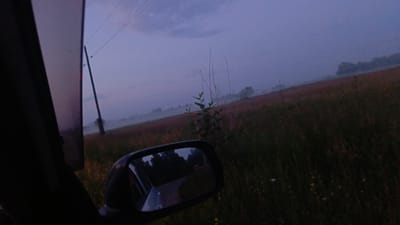 The image depicts a rural landscape at dusk, viewed from inside a vehicle. A side mirror reflects a road lined with trees. The sky is a muted purple and blue, with hints of clouds. A power line pole stands prominently in the background, suggesting the presence of infrastructure in the area. The terrain appears to be agricultural, with fields of grass and some scattered vegetation. The overall atmosphere is serene and slightly melancholic, possibly indicative of early morning or late evening hours. The image does not provide any specific visual cues to pinpoint its location within Angarsk, Russia.